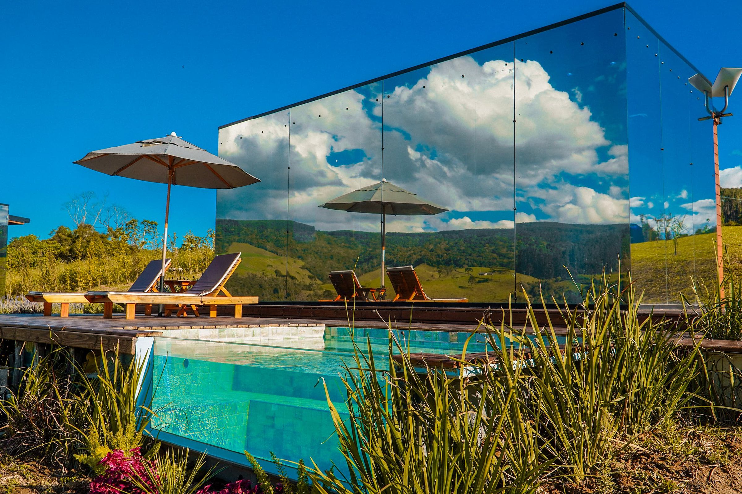 Cabana espelhada refletindo o céu e as montanhas, com piscina aquecida de vidro em primeiro plano